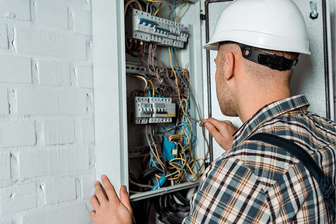 Electrician wearing a white hard hat inspects and repairs wiring inside an open electrical panel mounted on a brick wall.