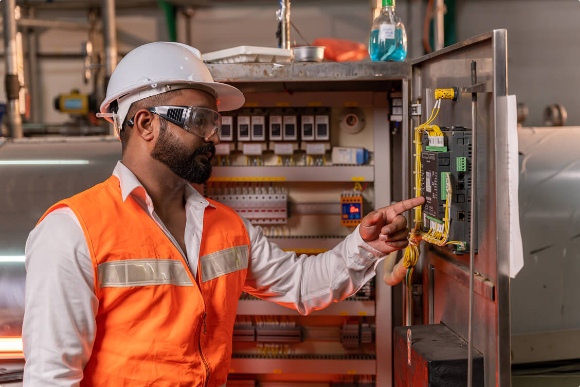 Industrial electrician in a white hard hat and orange safety vest inspects wiring and control systems inside an open electrical panel.