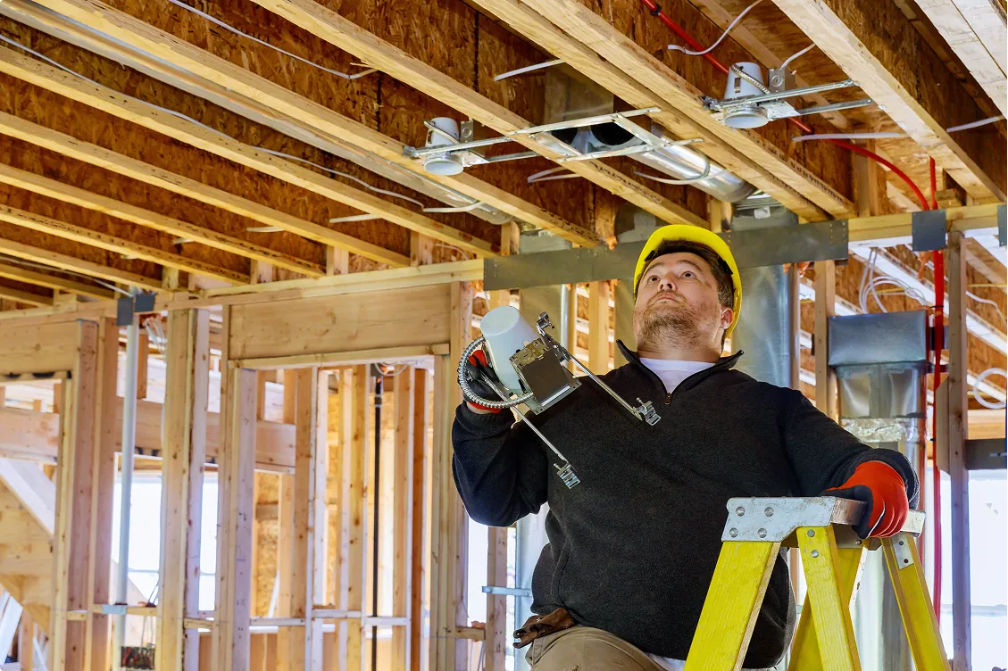 Construction electrician in a yellow hard hat stands on a ladder preparing to install recessed ceiling lighting inside a wooden-framed building.