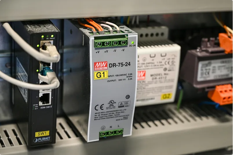 Close-up of an industrial electrical control panel featuring a Mean Well DR-75-24 power supply unit and network switch with connected wiring.