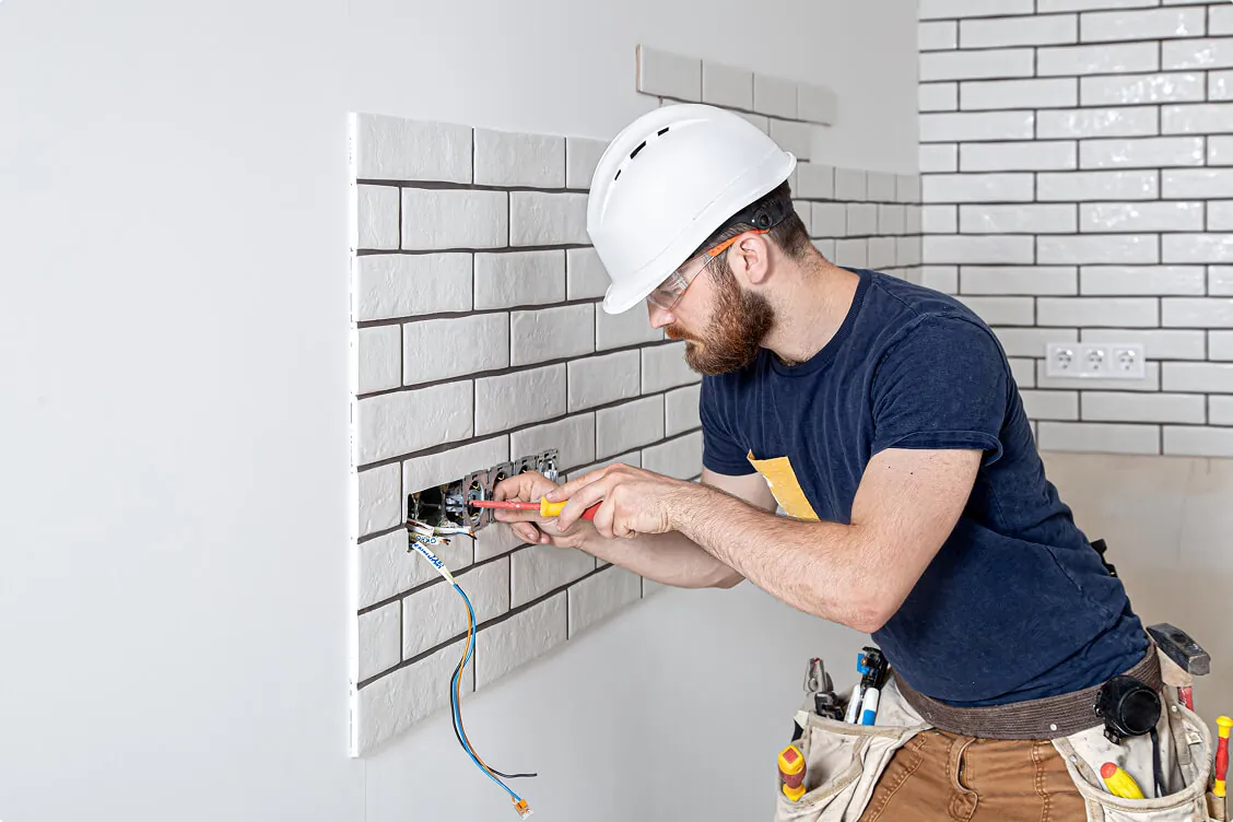 Residential electrician in a hard hat and tool belt installs electrical wiring inside a wall socket during home renovation.