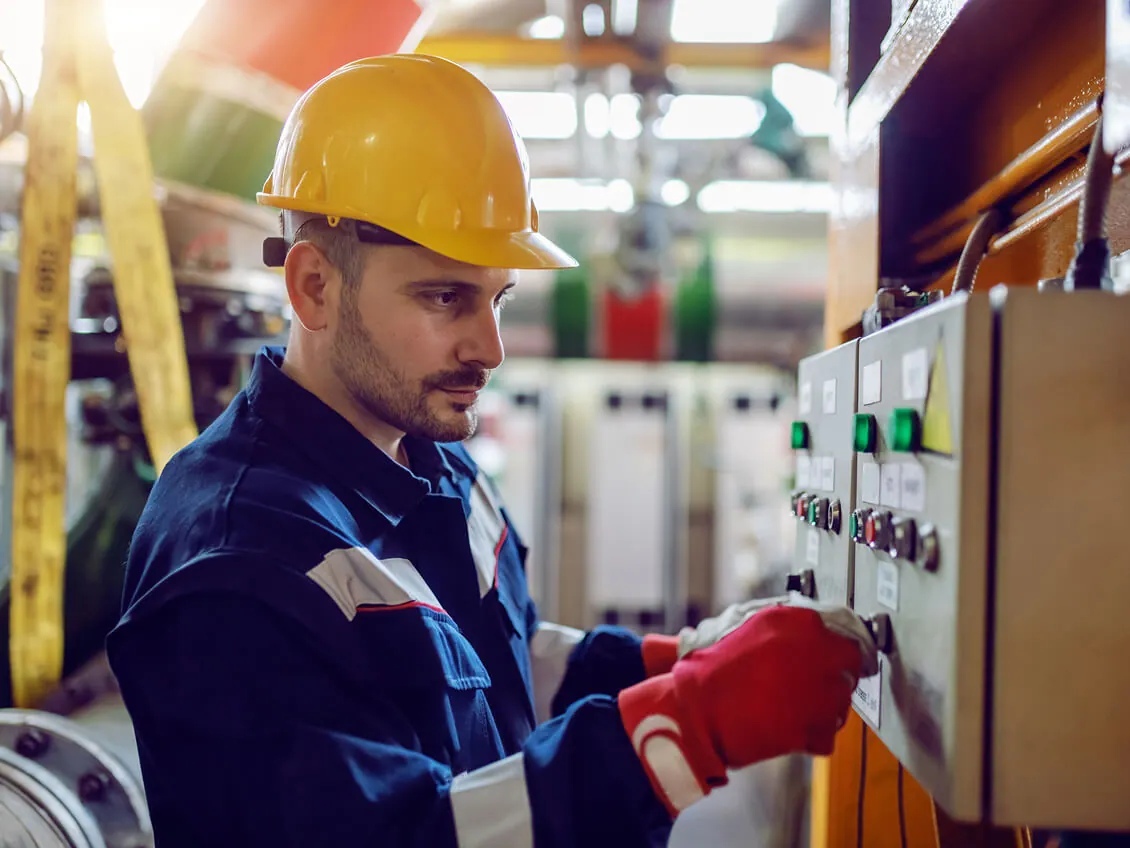 A technician wearing a hard hat and safety gloves operates control panel switches in an industrial electrical room with machinery in the background.