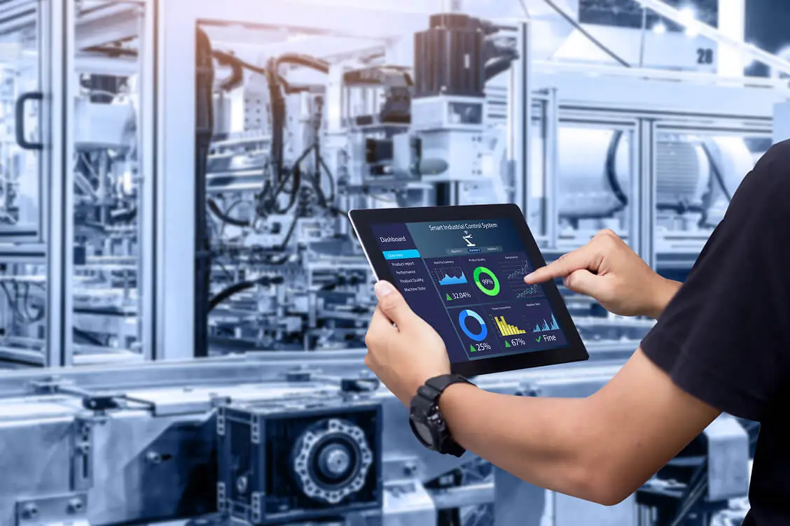A technician in an industrial setting uses a touchscreen tablet displaying performance dashboards and analytics graphs against the background of automated factory machinery.